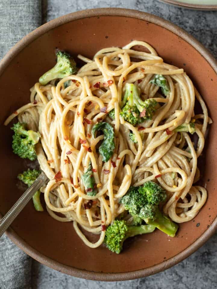 Overhead view of a dark red bowl filled with creamy tahini noodles and broccoli.