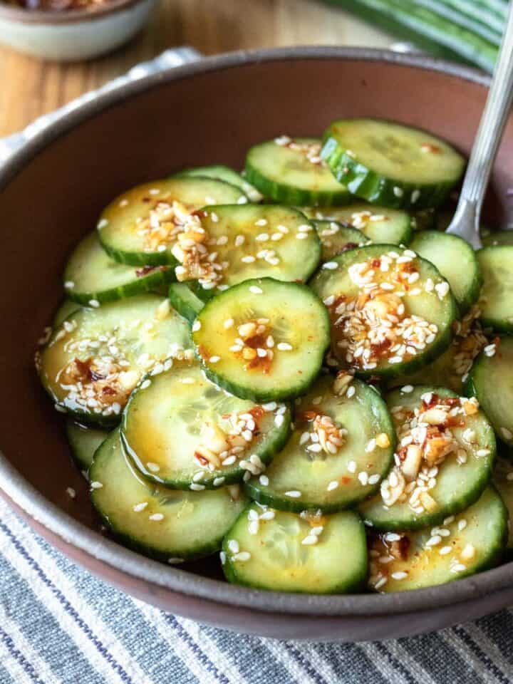 A dark red bowl filled with sliced cucumber tossed in spicy Asian dressing.